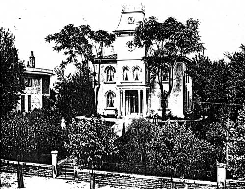 A large two-story home with a towering steeple-like structure on the front of it and tall trees and lush vegetation in the front yard