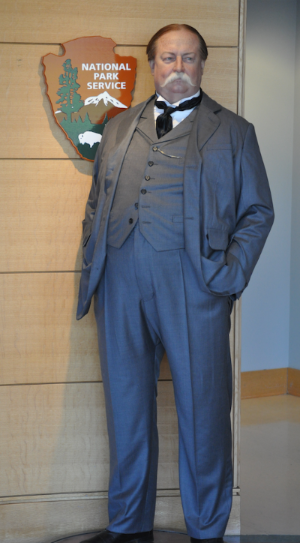 A wax statue of William Taft in a three piece suit wearing a wide handlebar mustache standing in front of the National Park Service arrowhead