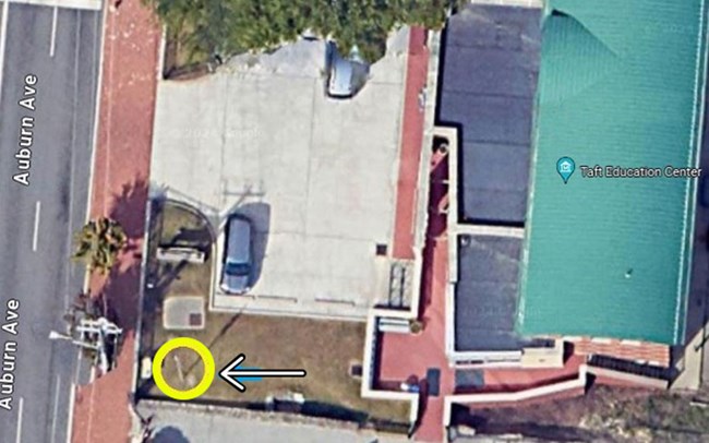 Aerial view of a building with a green roof on the right and a yellow circle on the lower left with a black and white arrow pointing to it, from the right side of the yellow circle