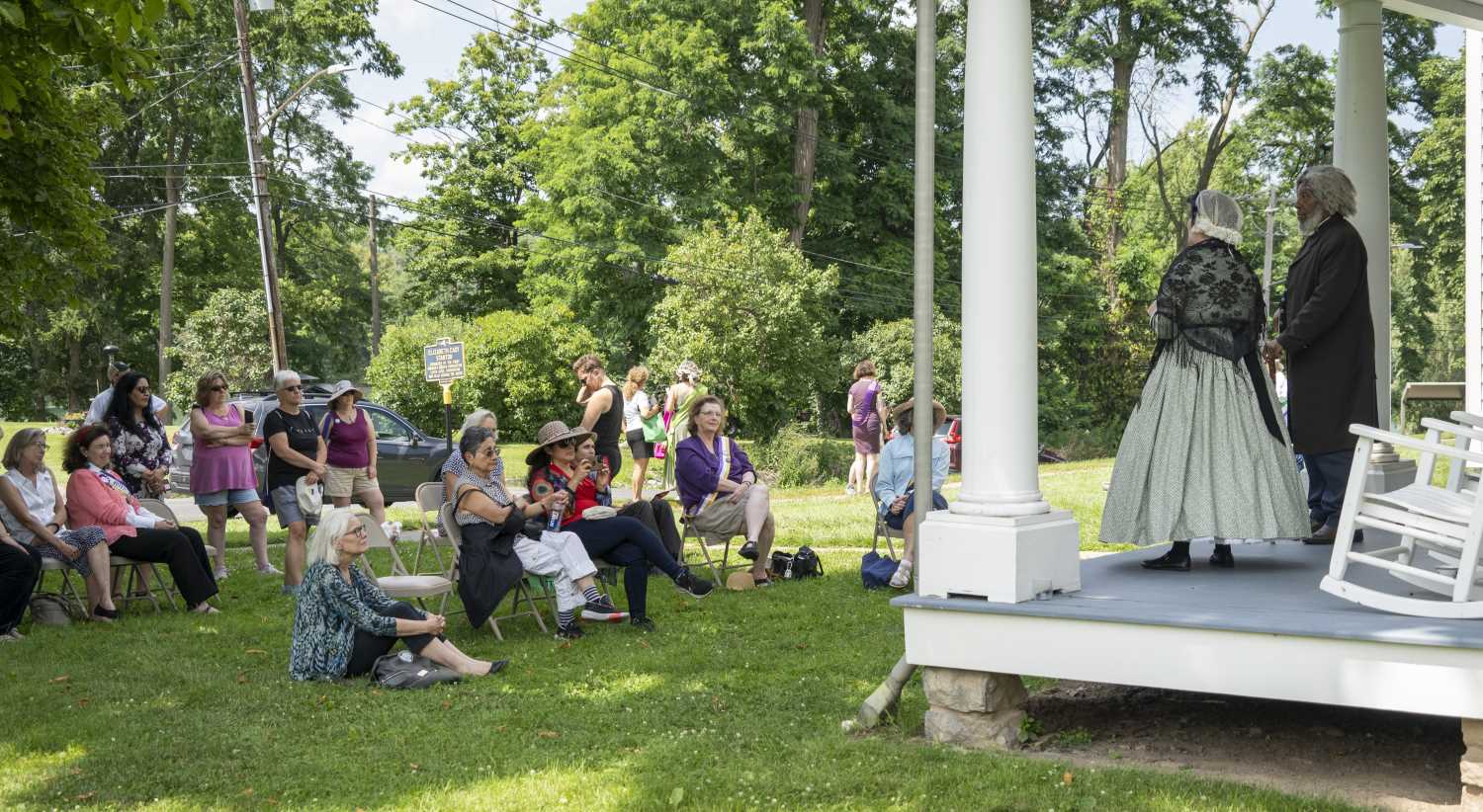 A man and women in historical clothing speak to a group of people from a porch.