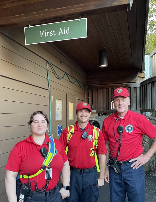 Three EMS volunteers stand under a sign that reads First Aid.