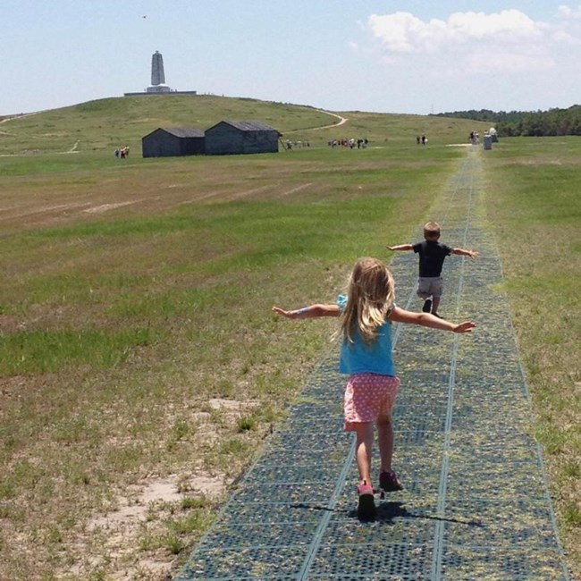 Two kids run down a path with arms outstretched like airplanes. Wooden buildings and the Wright Brothers monument are in the distance.