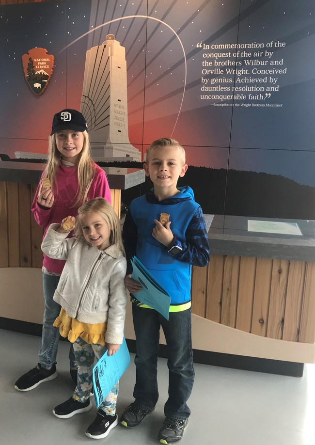 Three young children stand in front of a front desk proudly wearing their junior ranger badges. An image of the Wright Memorial is pictured behind them.