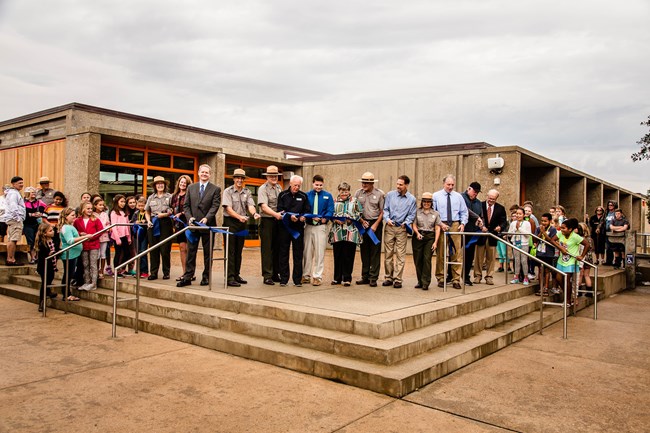 Group of adults and children stand on steps of visitor center holding a ribbon and smiling.