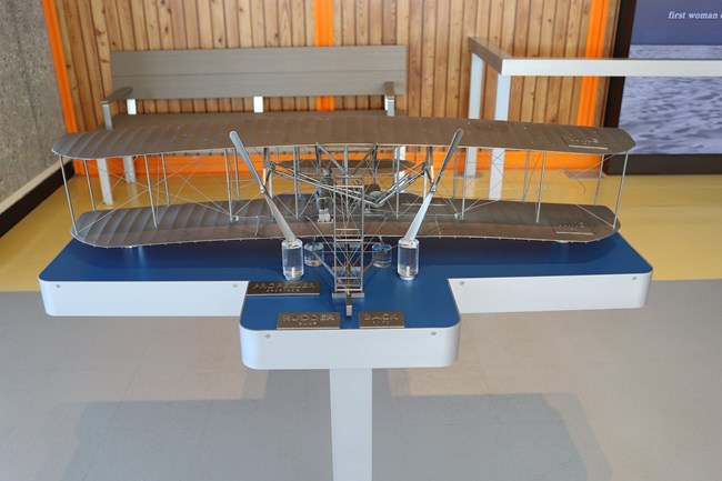 Silver model of an early airplane sitting on a base designed for easily reaching the model
