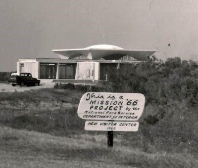 Black and white photo of visitor center with 1960s truck parked at it. Sign in front notes the building is a Mission 66 Project.