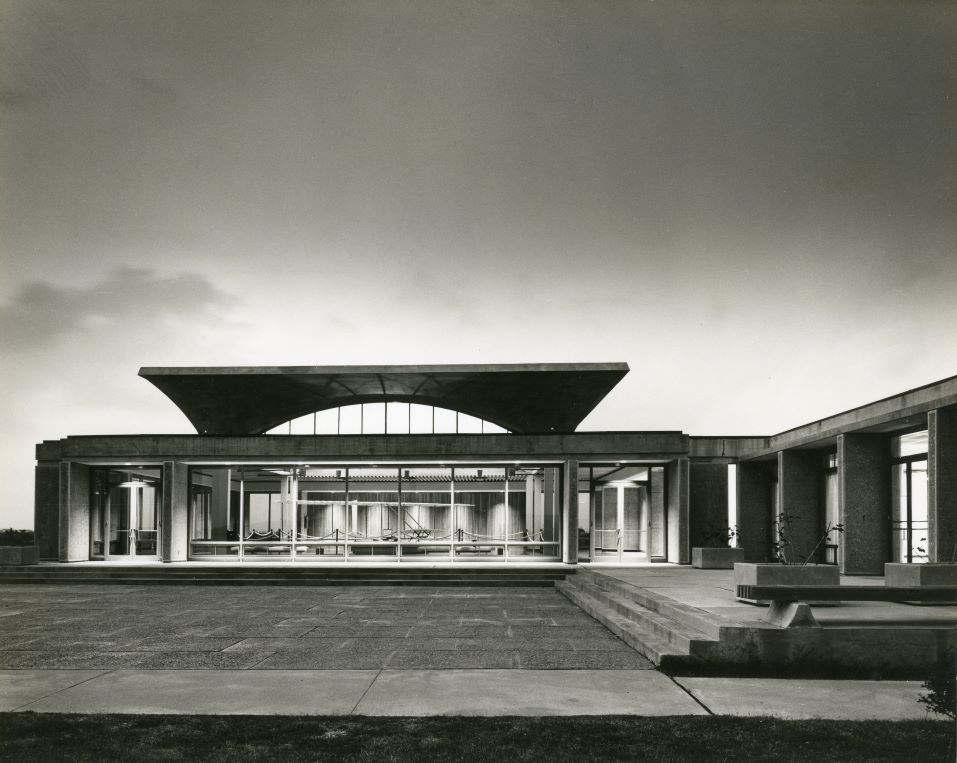 Black and white photo of building at twilight. Interior of building is seen through windows.