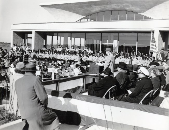 Black and white photo of dedication ceremony. Man stands at podium with crowd around him. Visitor Center seen in background.