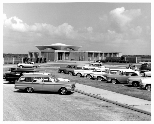 Black and white photo of parking lot with the Wright Brothers visitor center in background. 1960s-era cars seen parked.