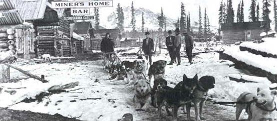 Historic photo of dog sled team with people in small town with wood buildings.