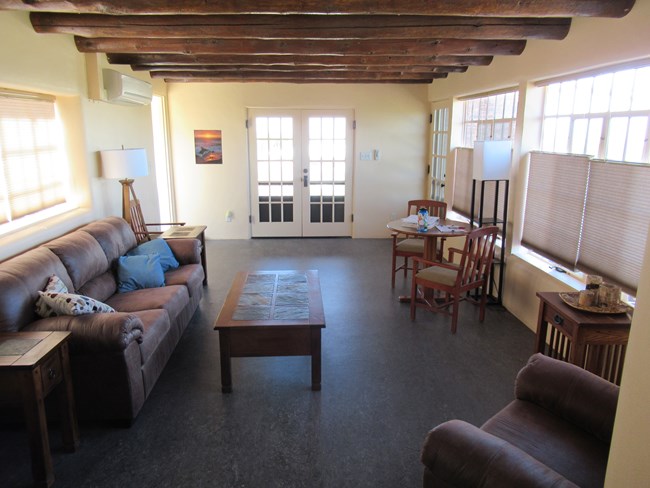 An interior living room with off-white plaster walls and a gray floor. There is a French style double door on the far wall, exposed wooden vigas in the ceiling, and large casement windows on either side.