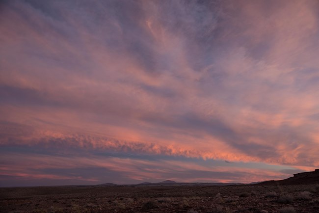 An open landscape of sandstone mesas and dry grasses with a pink and light purple sky and wispy clouds.