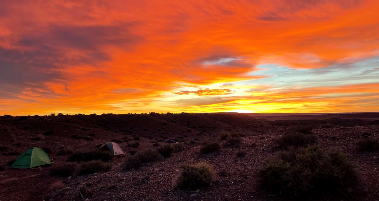 Two domed camping tents set up among black cinders and red sandstone with a bright yellow and red sunrise in the background.