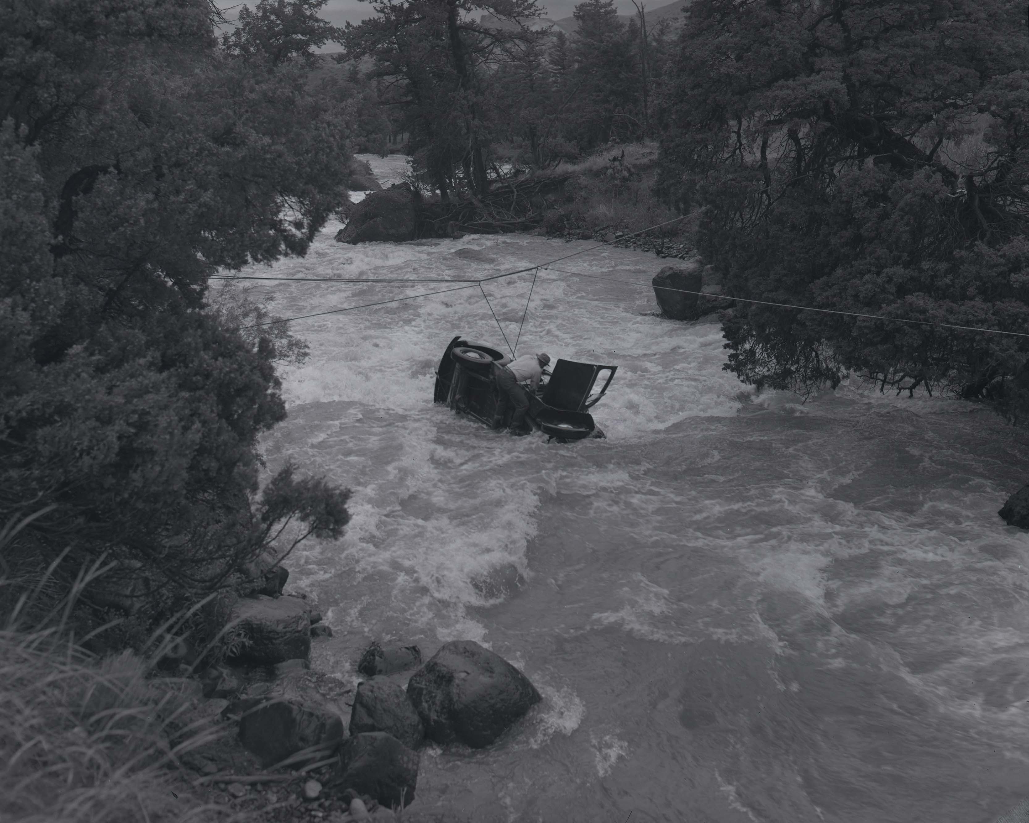 Image of car on its side in the middle oa a river, attached to wires. A man stands in the river bent over the car.