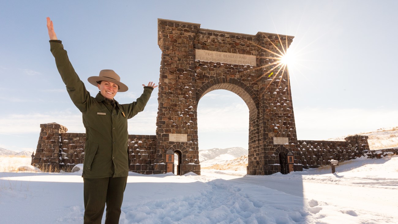 A park ranger in uniform standing near a large stone archway with their hands outstretched above their head