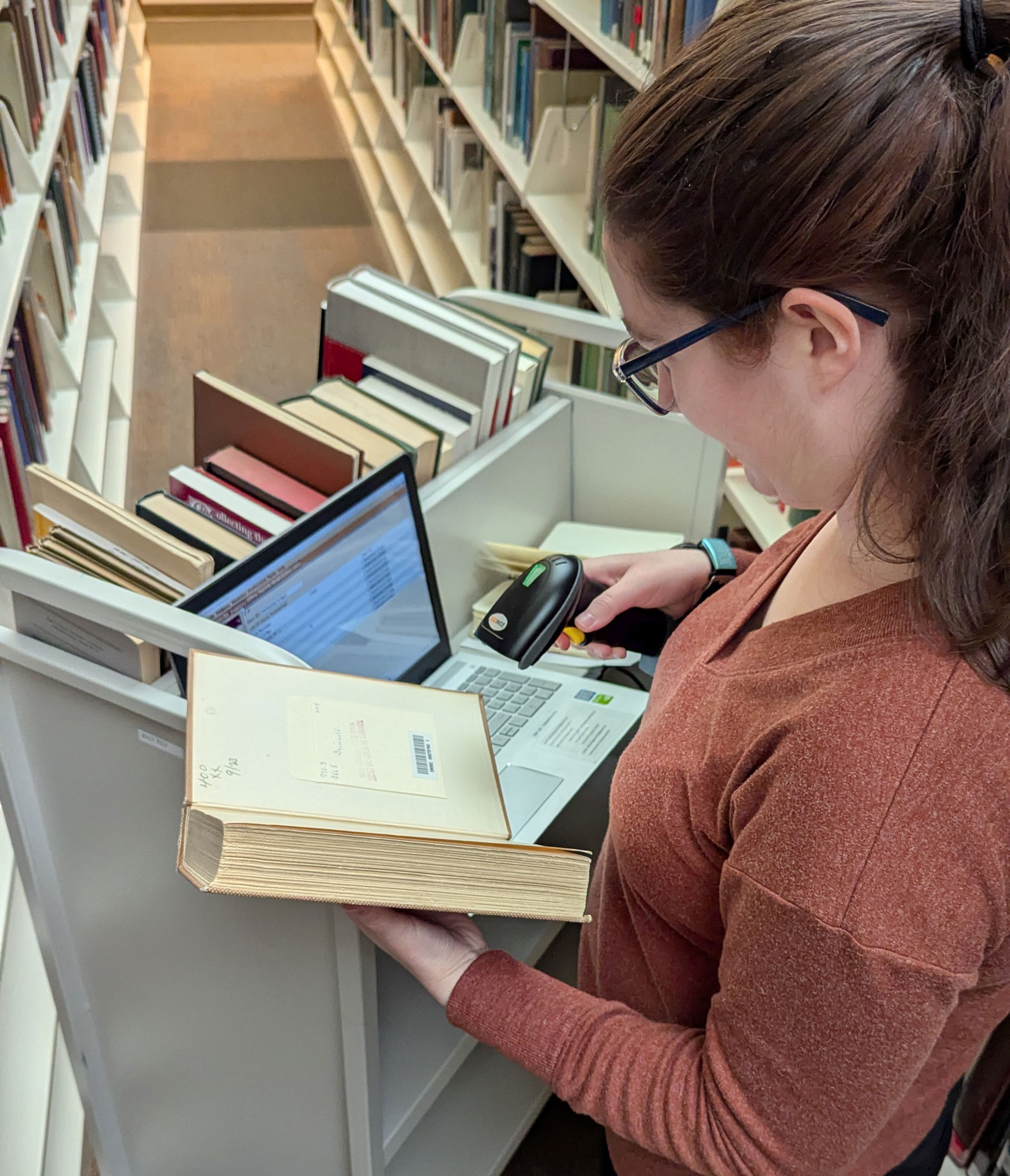 Photo of woman in red sweater scanning an open book with laptop in the background.