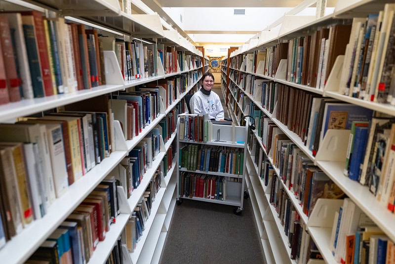 photo of woman in white sweater sitting behind a cart of books in between two bookshelves.