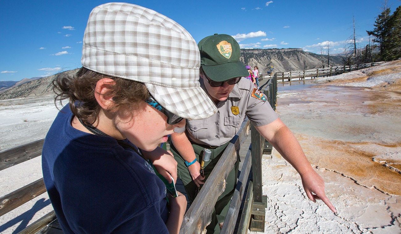 A ranger on a boardwalk points to something as a student looks on. There is a hot spring in the background.