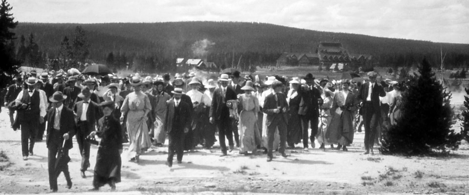 A historic photo of a large group of visitors walking away from the Old Faithful Inn.