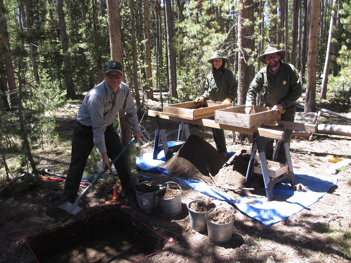 Three uniformed people use a shovel and boxes to excavate an archeology site.
