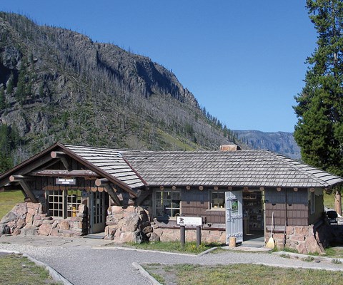 A one-story stone and log building with wooden shingles beneath a mountain