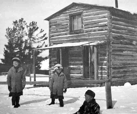 Three children in winter clothes stand in front of a two-story log cabin