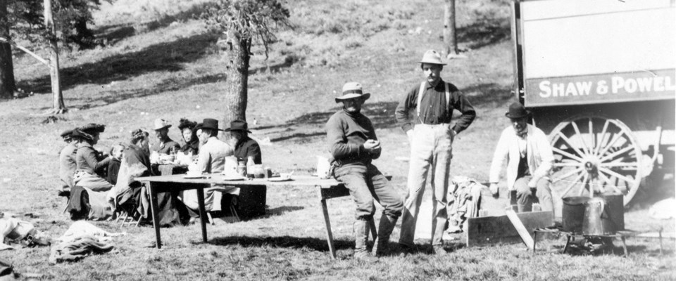 A black and white photo of people sitting at a table eating and three men resting next to a wagon