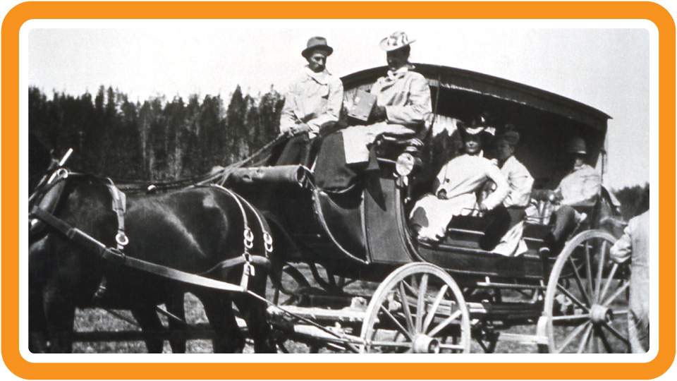 A man and woman sit at the front of a stagecoach, holding the reins, while three other passengers look out from the coach.