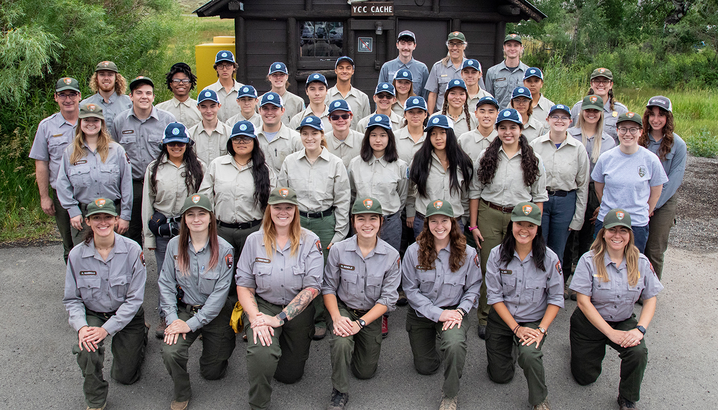 A group of teens in tan uniforms and adults in green and grey uniforms pose in front of a dark brown cabin.