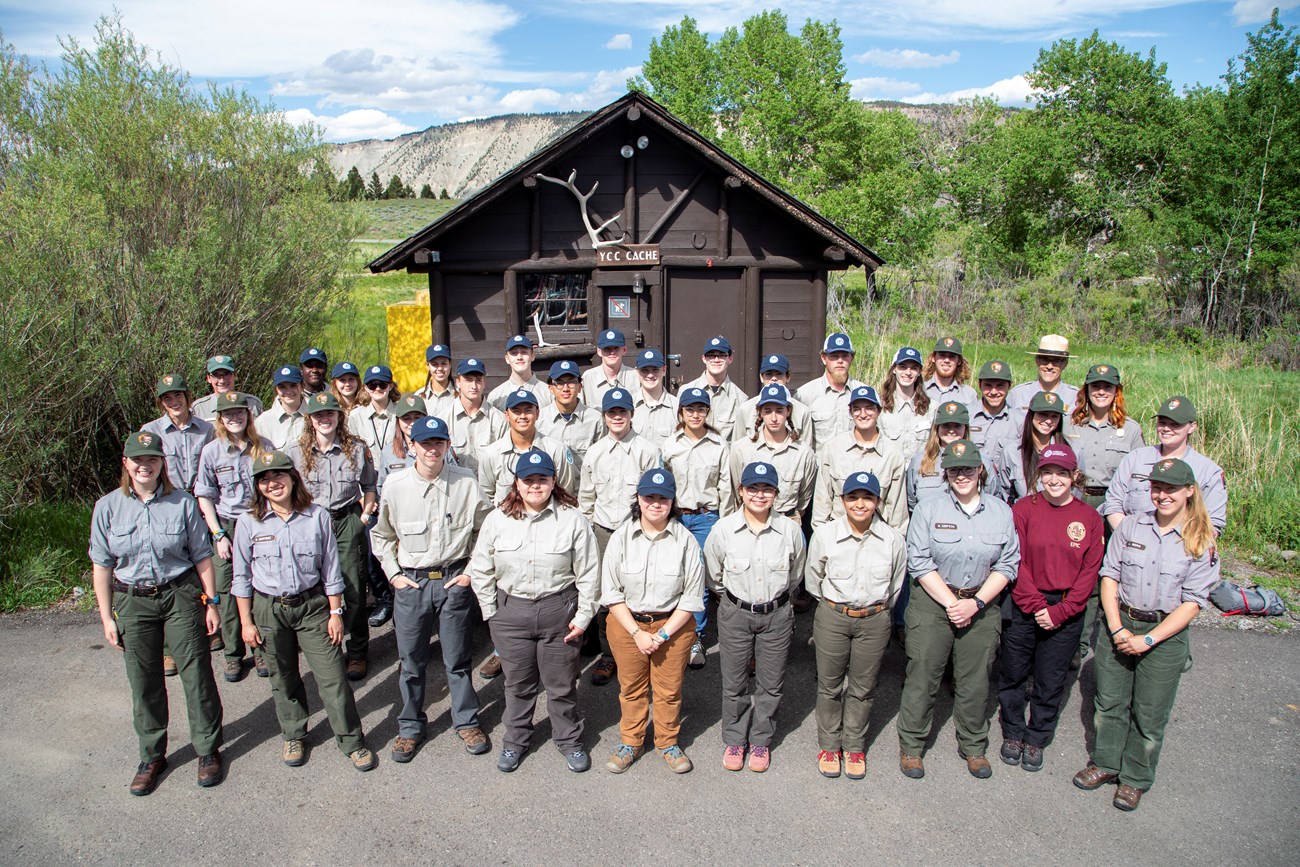 A group of teens in tan uniforms poses in front of a brown cabin in a brushy meadow with adults in green and grey uniforms.