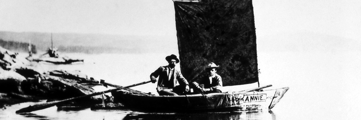 Two men sit on a wooden boat with a square sail