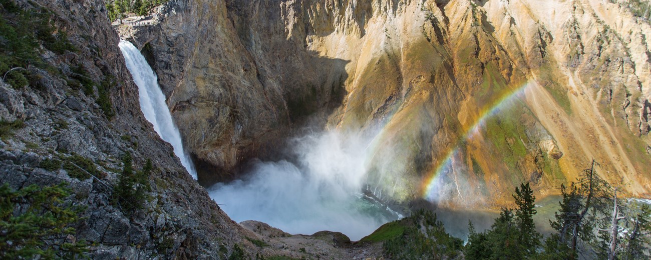 A waterfall is viewed from the side as it falls down into a steep, tan-walled canyon.