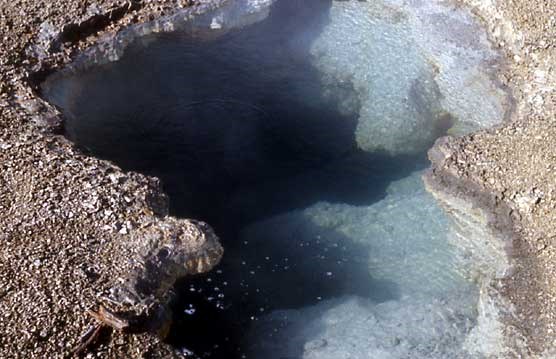 A large, wide hole in the rock is filled with hot, blue water, resembling a large mouth.