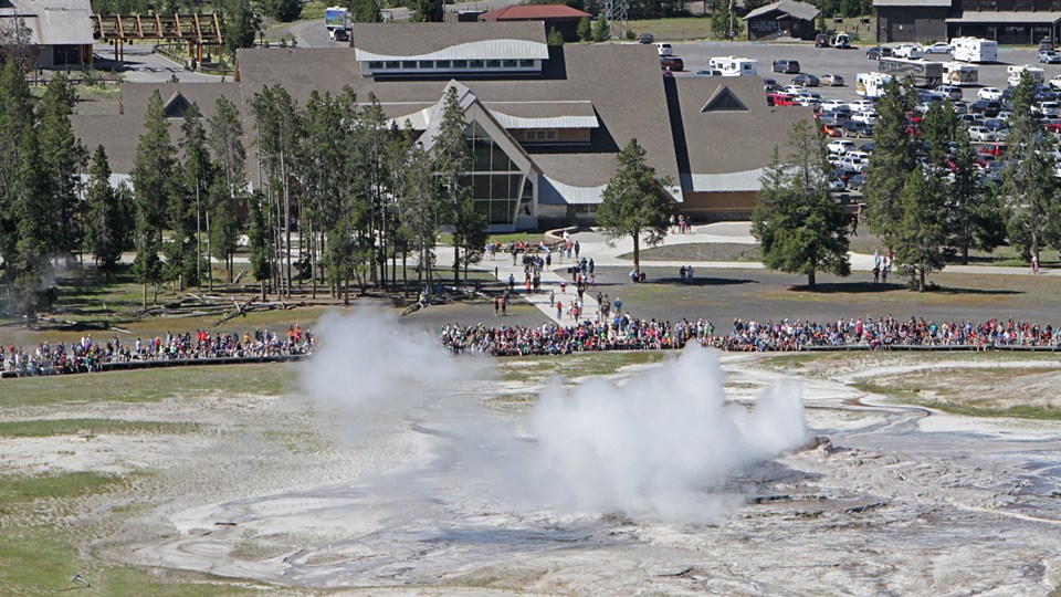 View of Old Faithful Geyser with visitors watching and the Old Faithful Visitor Education Center in the background