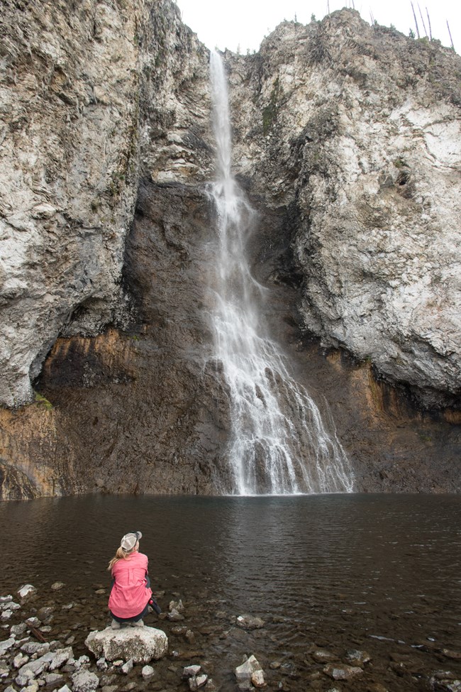 A hiker squats next to the pool at the base of the towering Fairy Falls.