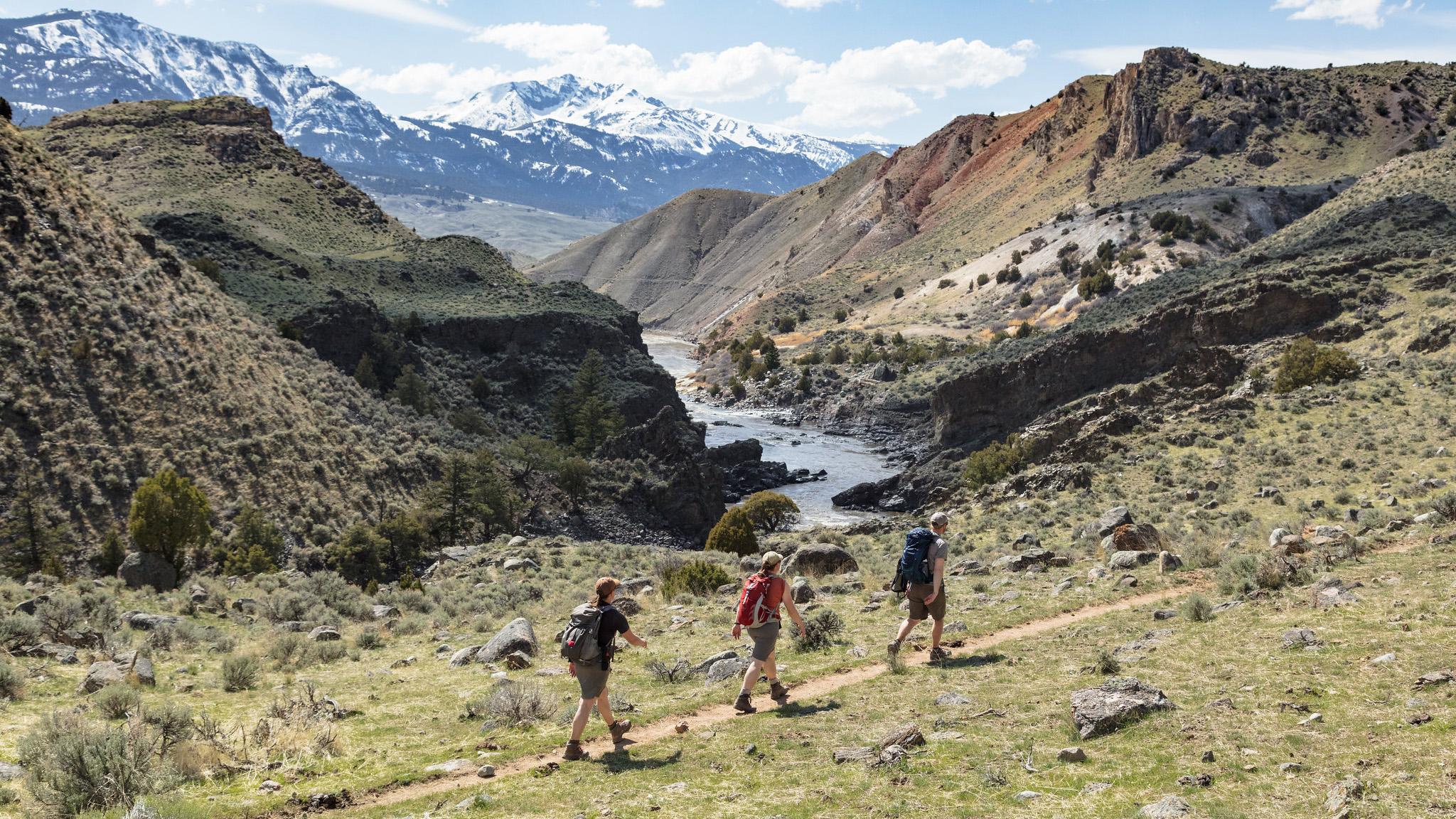 Hikers on a trail near a river and snowy mountains
