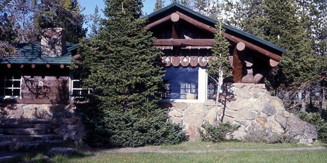 A rustic log building built on stone is surrounded by pine trees