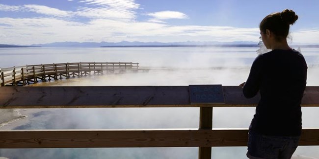 A young woman looks over a steaming pool and a lake from the railes of a boardwalk