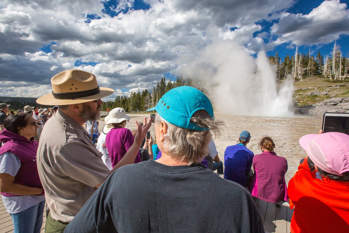 A ranger speaks to visitors in front of a geyser that's erupting