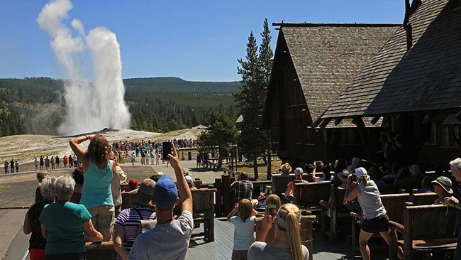 Crowd on an outdoor balcony watching a geyser erupt water and steam into the air.