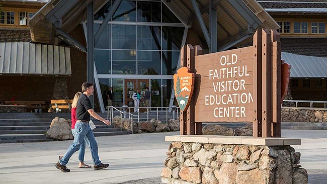 Couple walks in front of a tall, glass-paneled building and behind a sign that reads "Old Faithful Visitor Education Center".