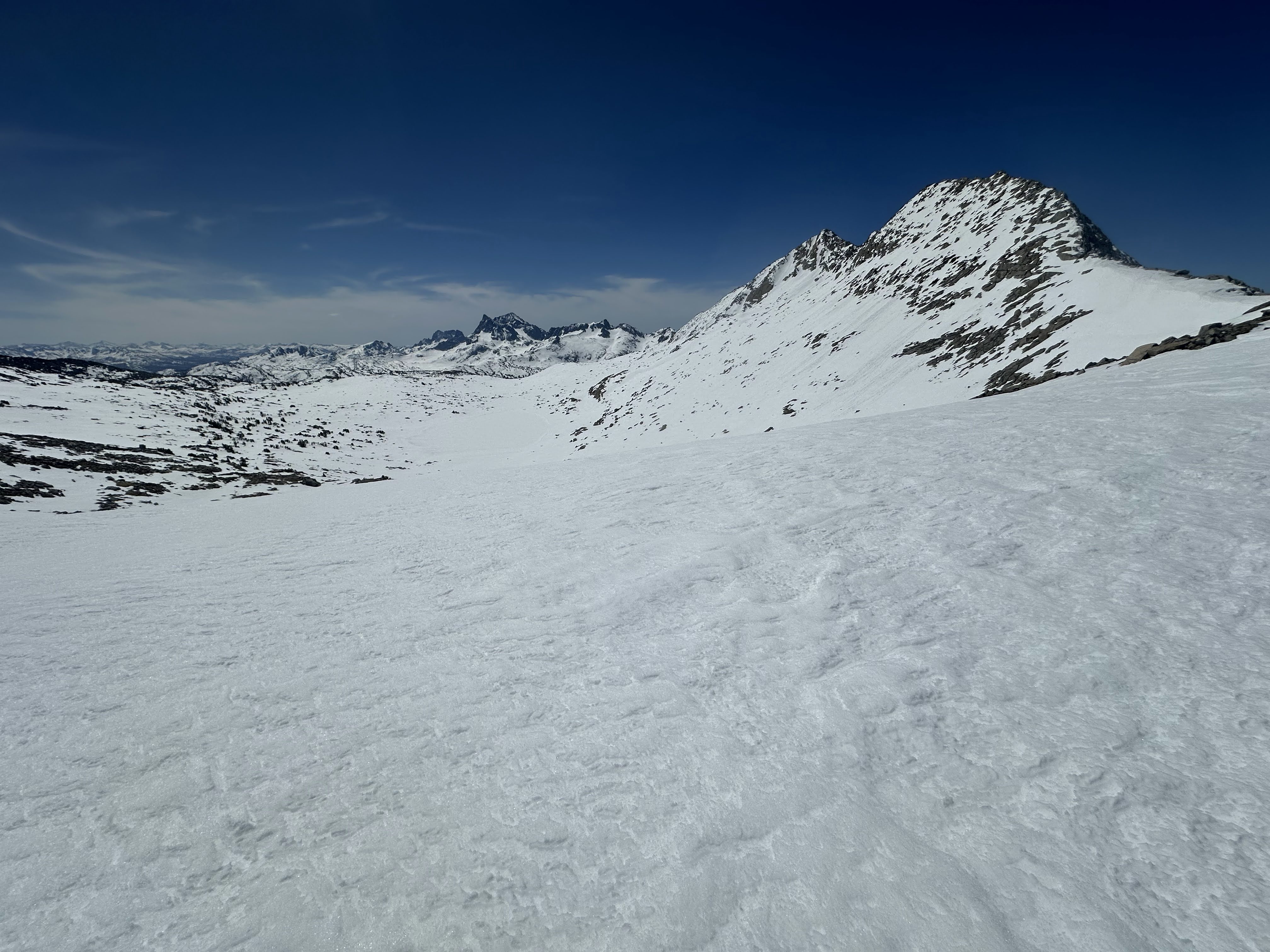 Standing on the snowy shoulder of a mountain peak with a snow-covered lake in the mid-distance and snowy peaks in the far distance