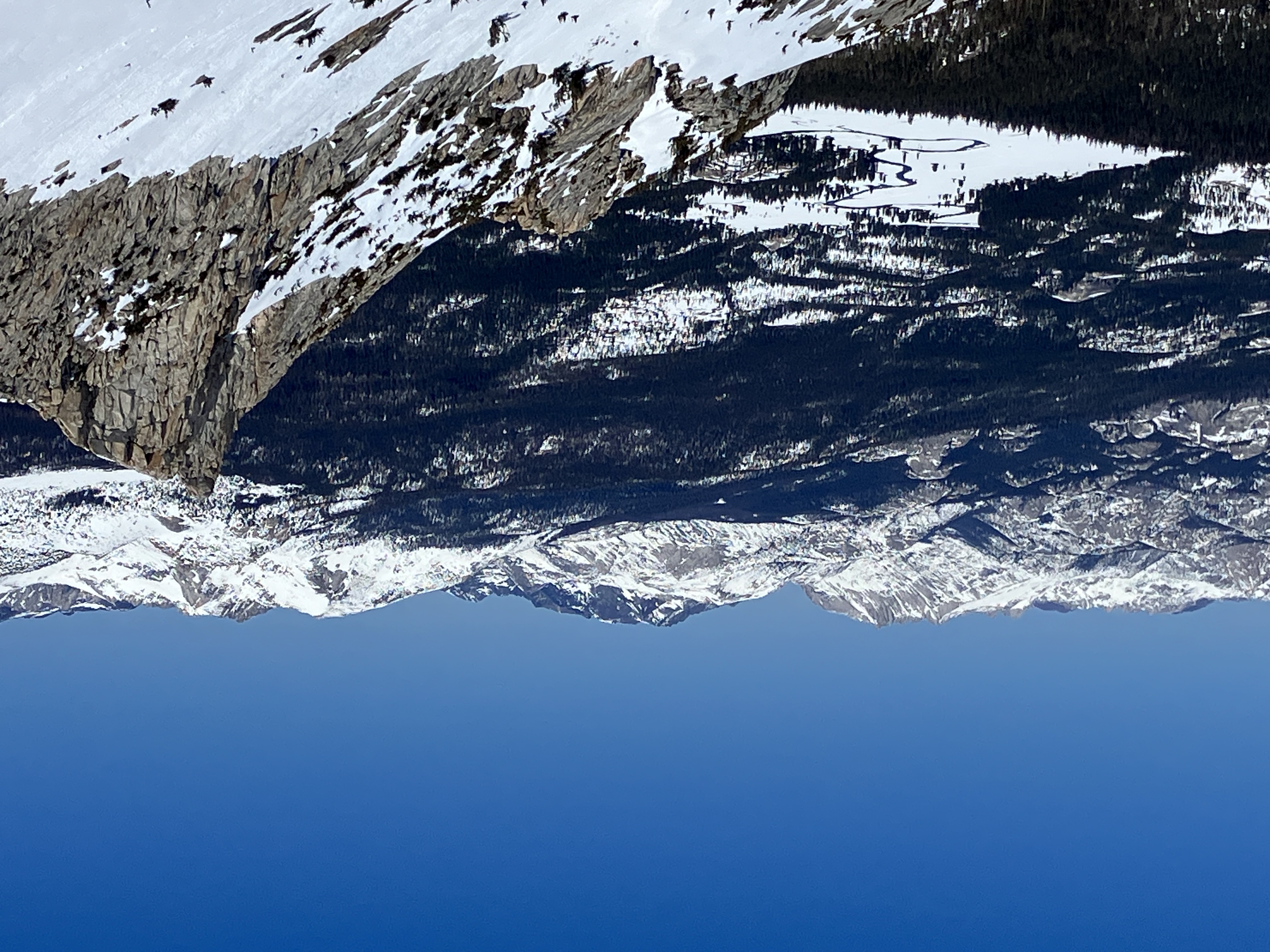 Snow-covered Tuolumne Meadows, with a river winding through it, looks small when looking from above and from a distance; it's surrounded by forest and partly snowy peaks