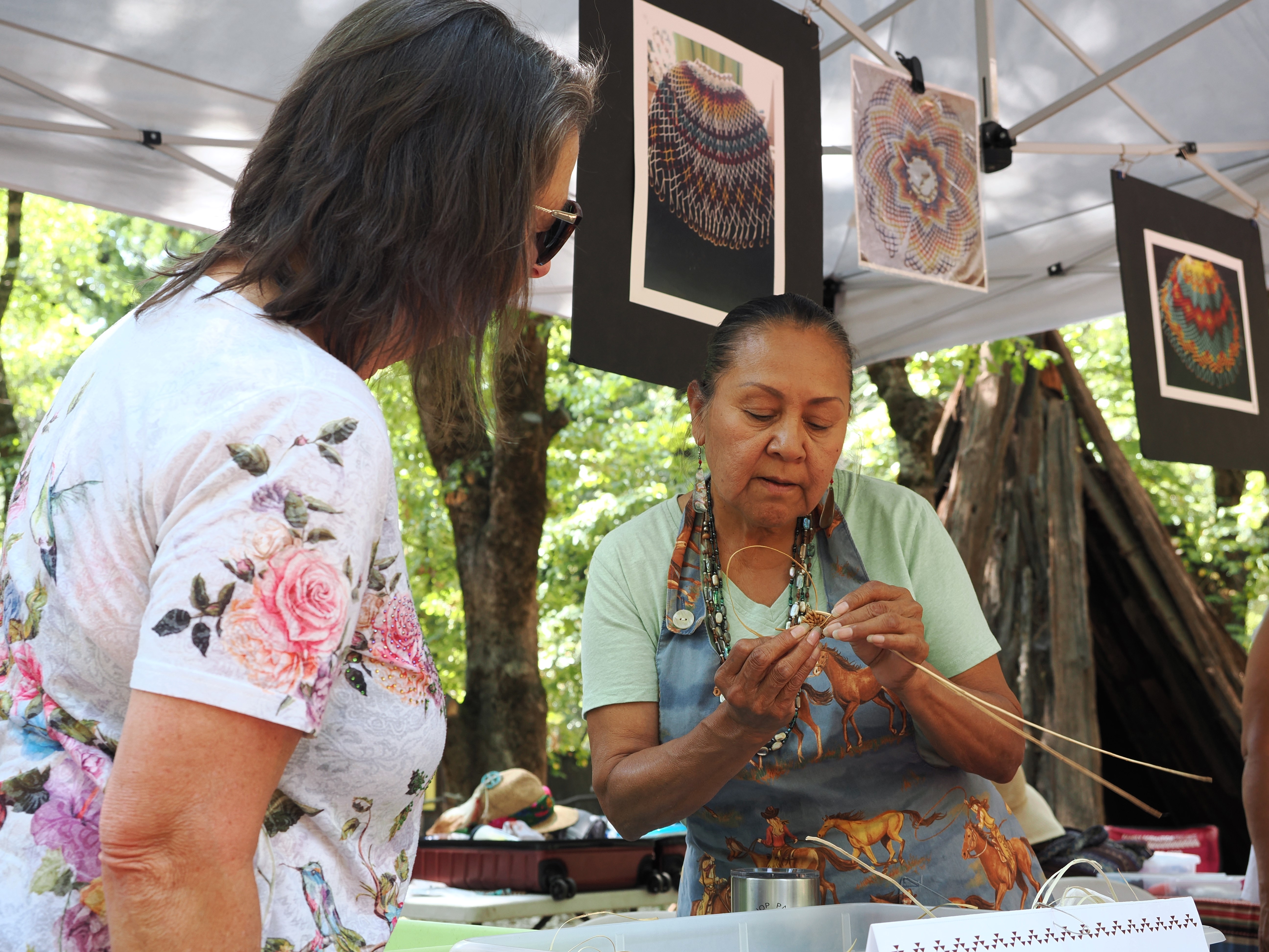A picture of two women talking closely. The person on the right is holding some strands of plant material woven into the beginning of a basket up for the other woman to look at.