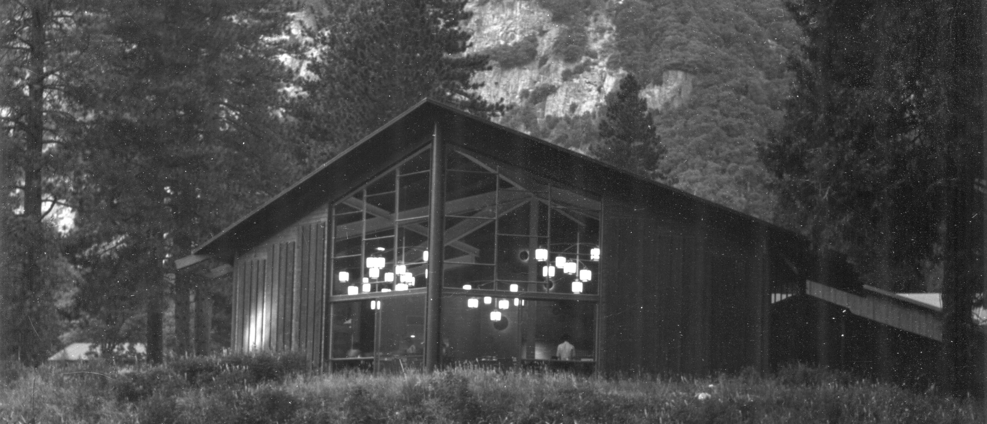 Grasses in front and cliffs behind frame a large window wall with a low angled roof, lights are visible inside