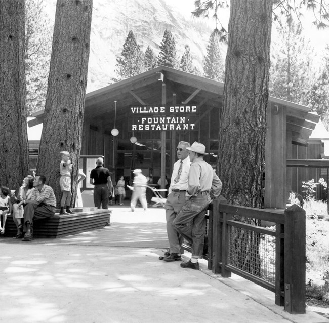 Park visitors stand and walk around a plaza in front of a store entrance; mature trees decorate the plaza
