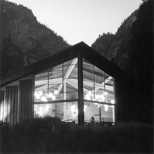 The window wall of a restaurant is shown brightly lit with asymmetrical chandeliers and exposed beams on the vaulted ceiling