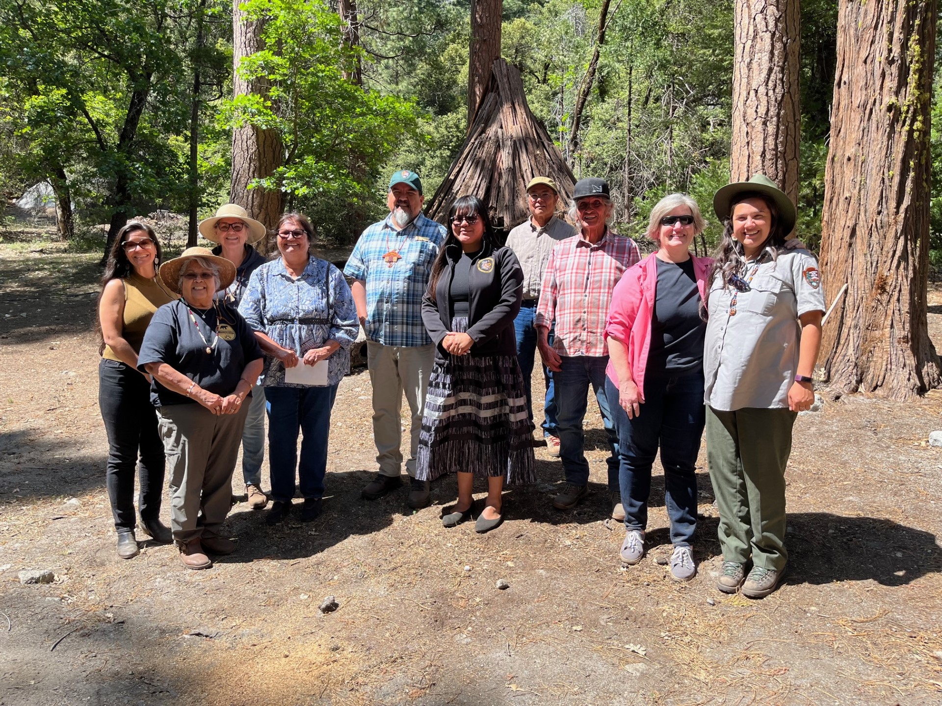 Ten people standing in front of an umacha, or traditional bark house, in a forested area.