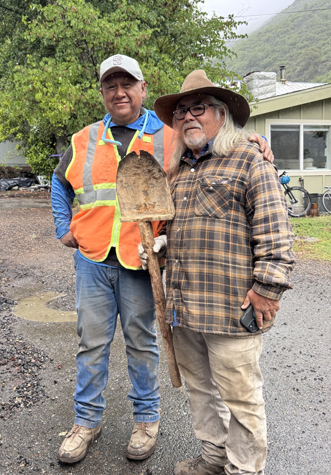 A picture of two men, one wearing a safety vest and the other holding a shovel, standing in front of a building in the mountains.