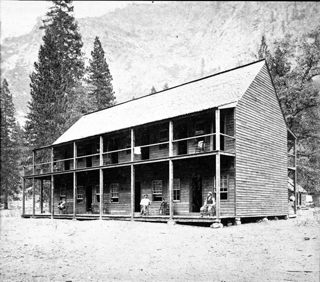 A two story wood building with porches on both levels. Two men sit on chairs on the porch.